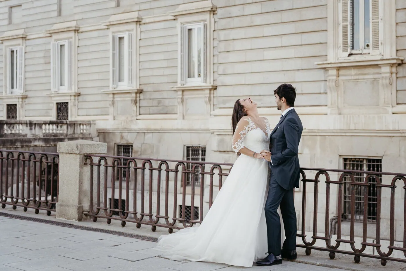 Pareja en atuendos nupciales sonríe y sostiene manos frente a edificio clásico.