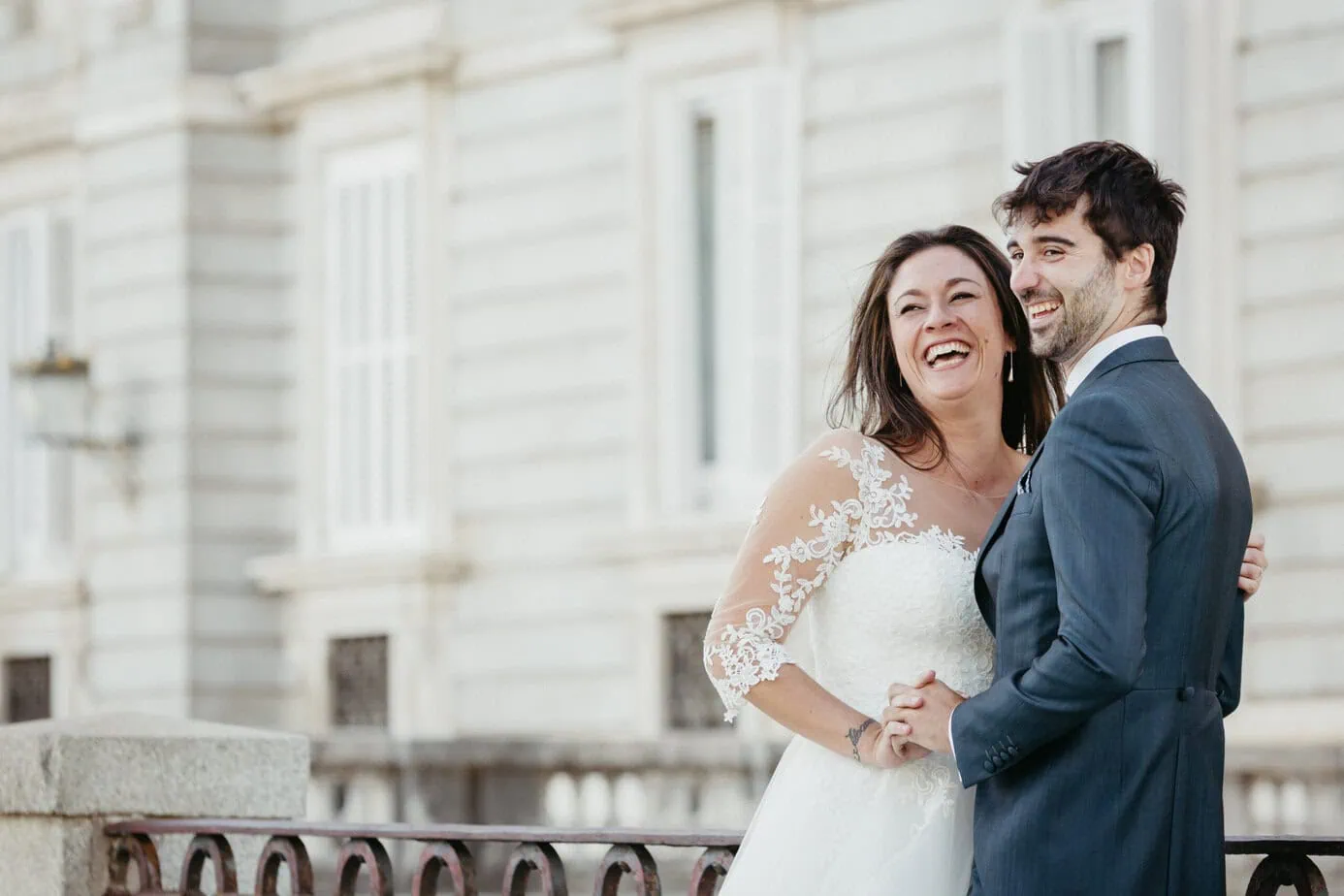 Pareja sonriente vestida de novios posa al aire libre frente a edificio clásico.