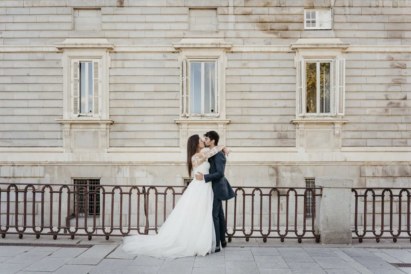 Pareja de novios besándose frente a un edificio de piedra con ventanas clásicas.