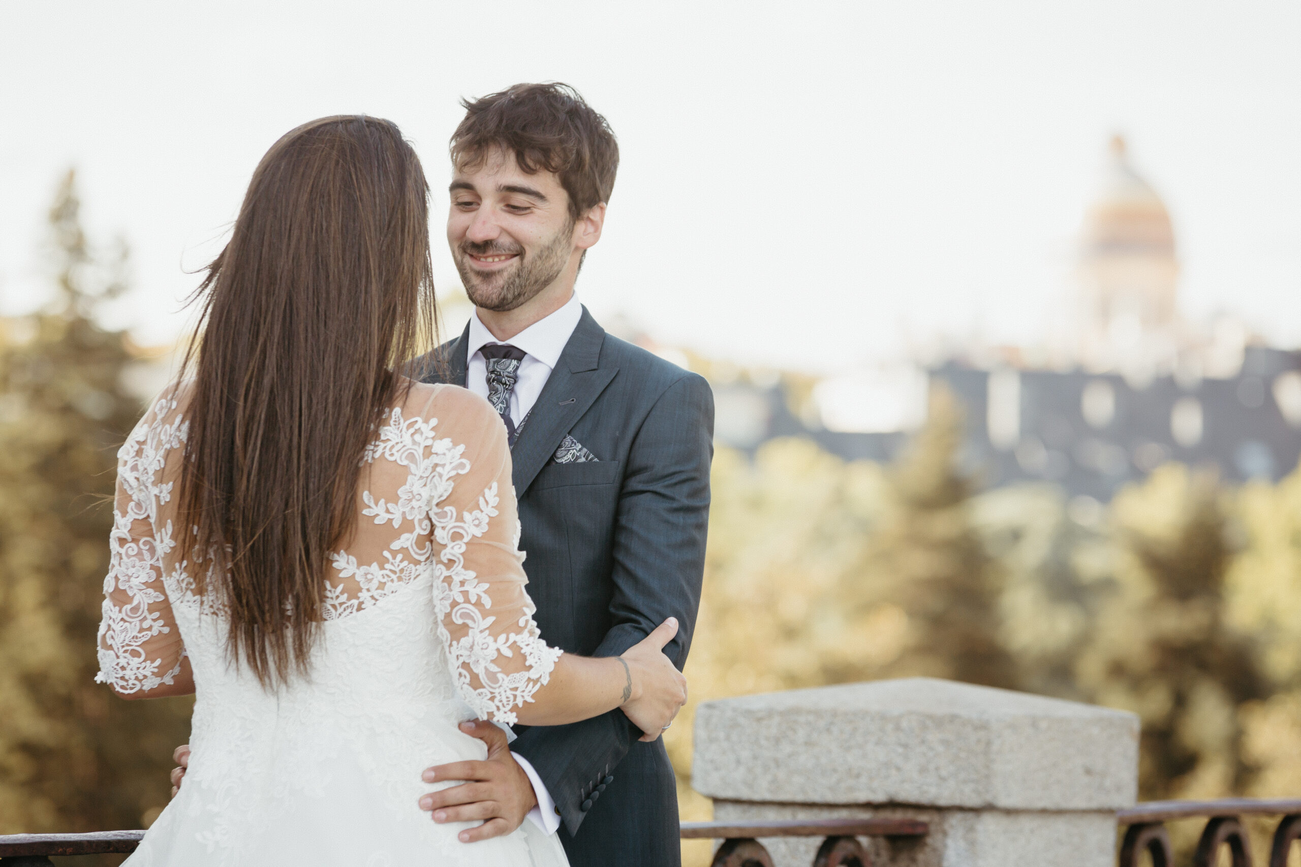 Pareja vestida de novios sonríe al aire libre, con fondo urbano difuminado.