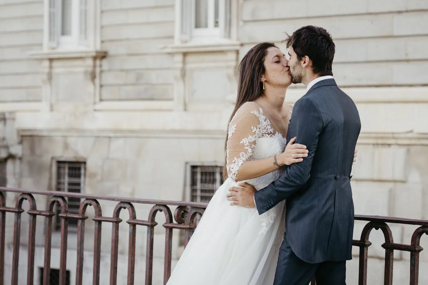Pareja besándose frente a un edificio de piedra junto a una barandilla de hierro.