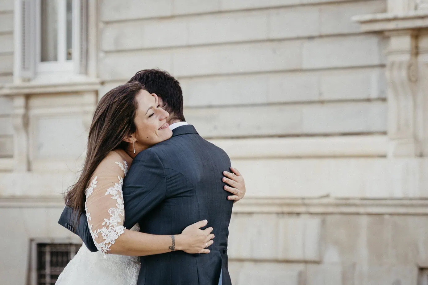Pareja abrazándose frente a un edificio de piedra, ella sonríe en vestido con encaje.