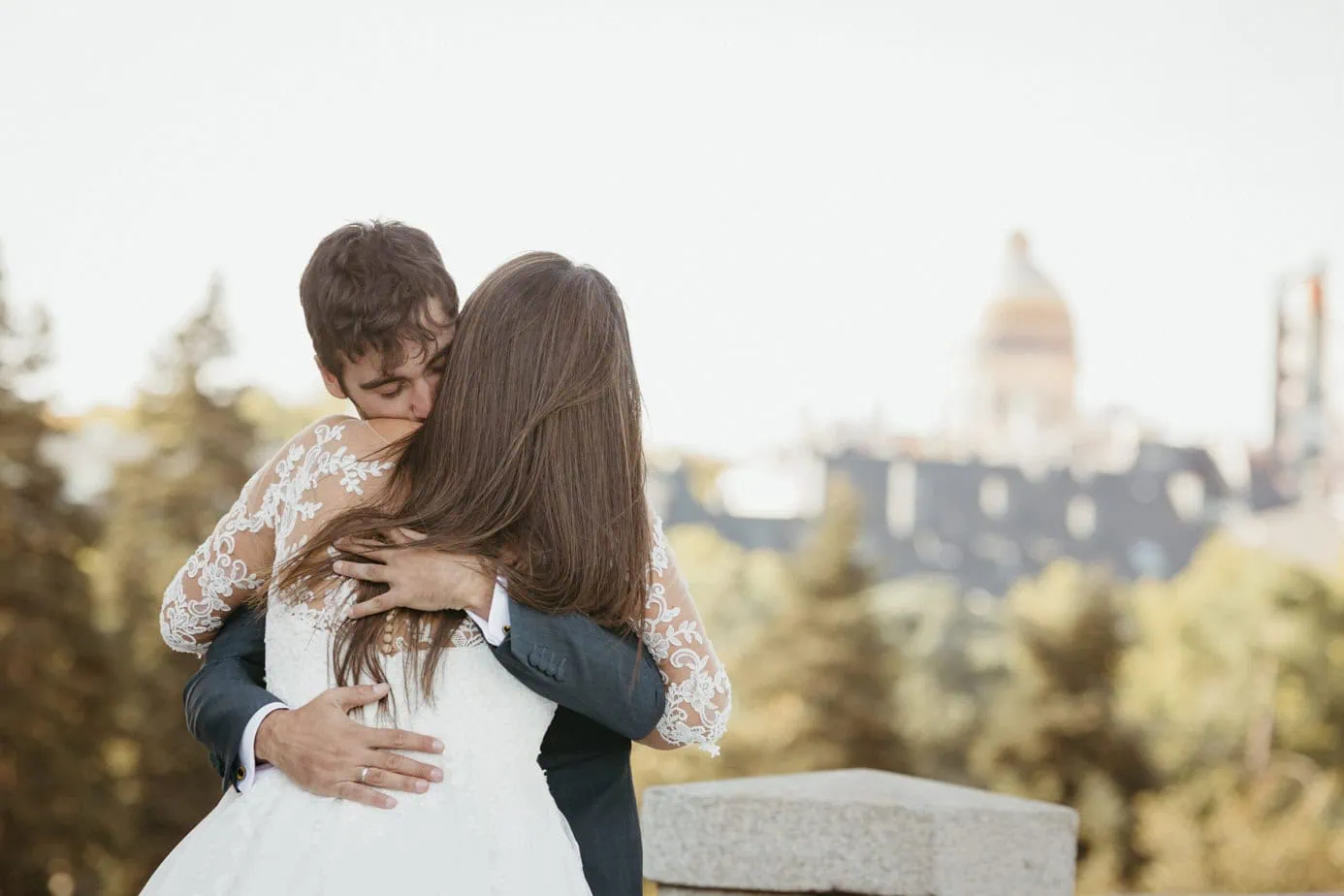 Pareja en traje y vestido blanco se abraza con un edificio de cúpula difuminado al fondo.