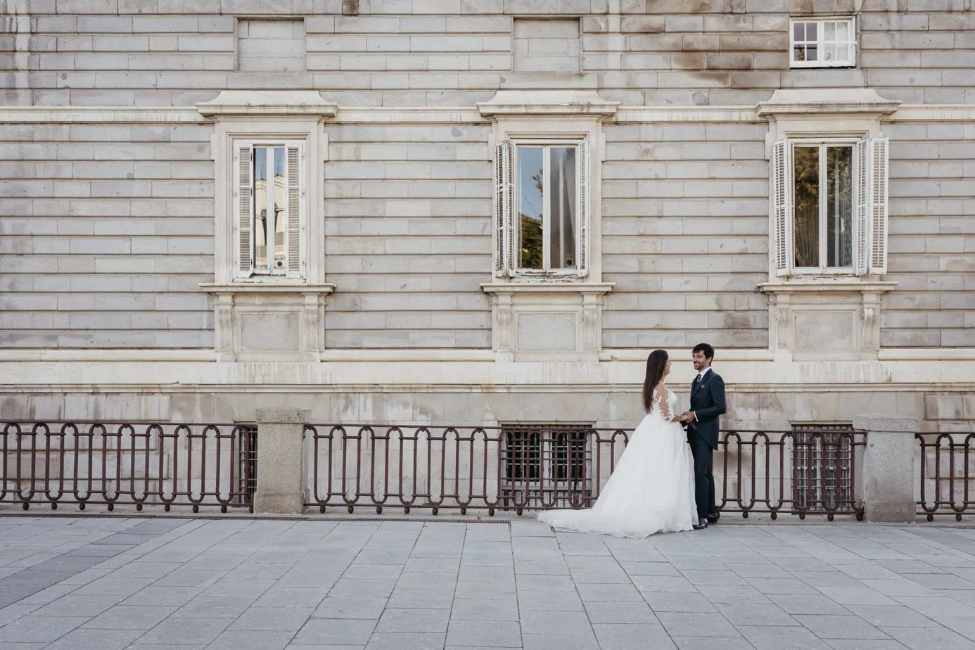 Pareja de novios frente a edificio de piedra con ventanas grandes en zona urbana pavimentada.