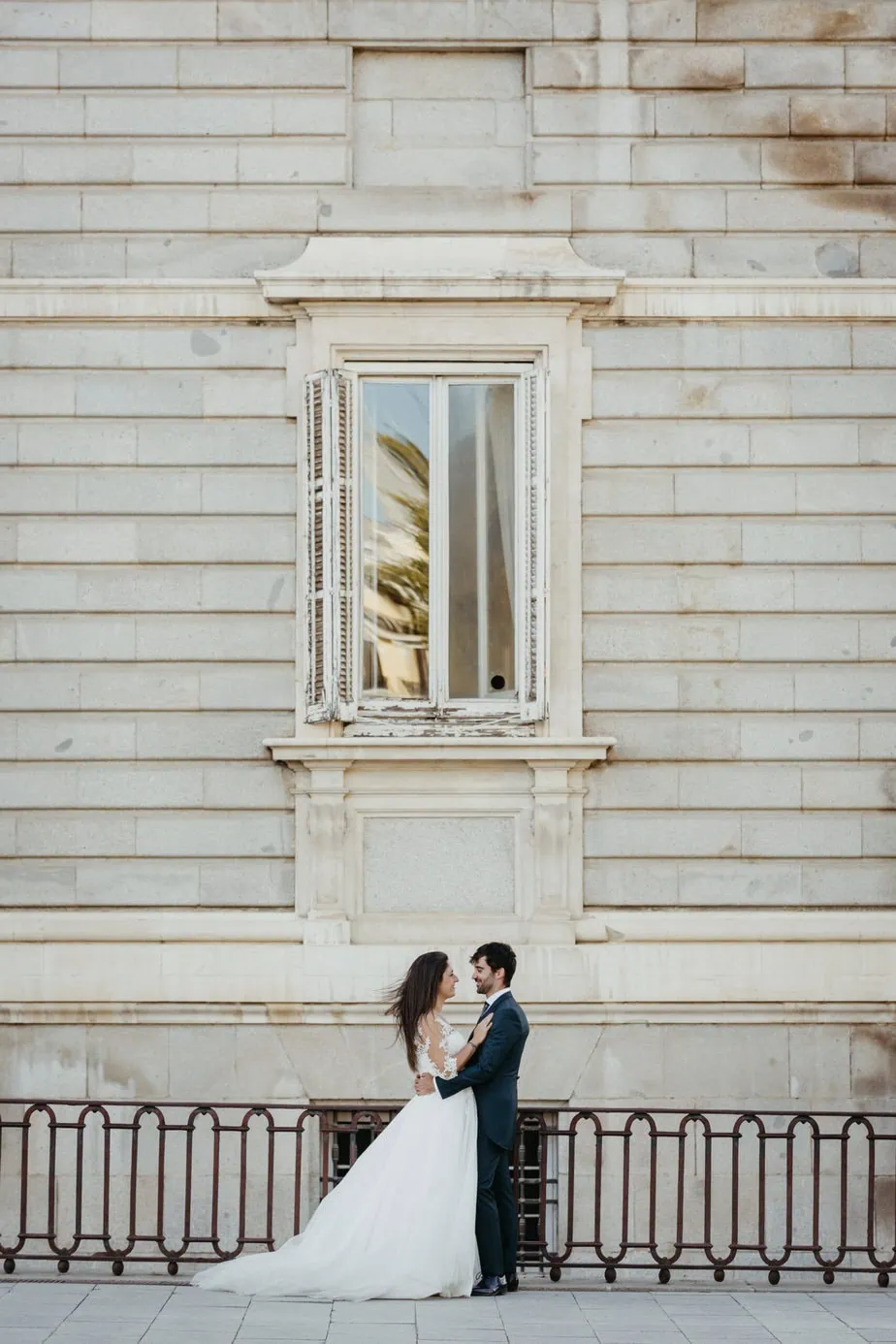 Pareja elegante abrazándose frente a edificio con ventana abierta, vestido blanco y traje oscuro.