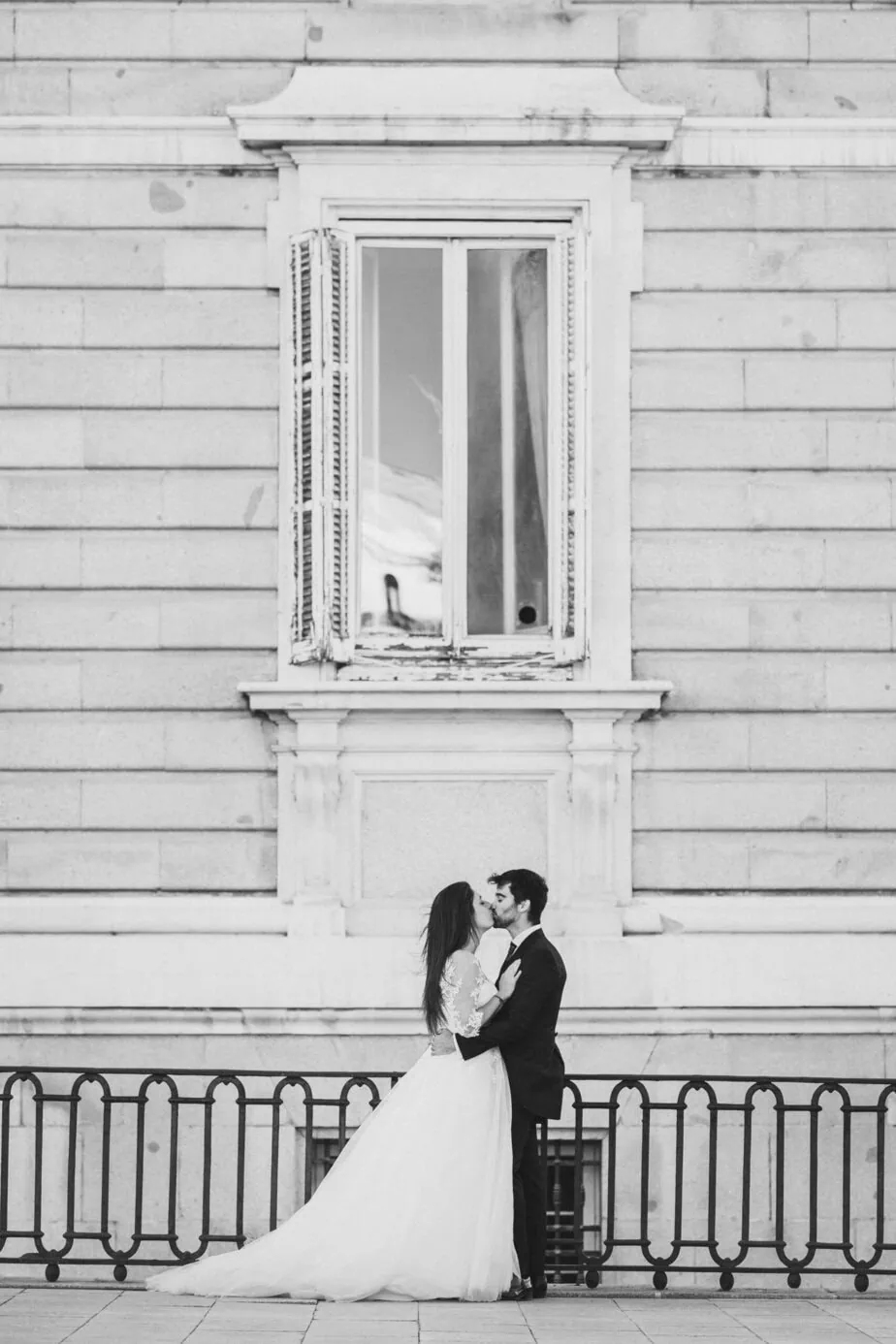 Pareja de boda besándose frente a muro de piedra clara y ventana en blanco y negro.