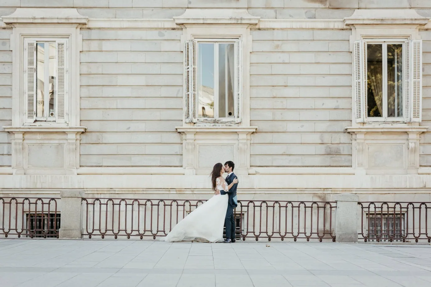 Pareja en bodas besándose frente a edificio de piedra con ventanas grandes y barandilla.