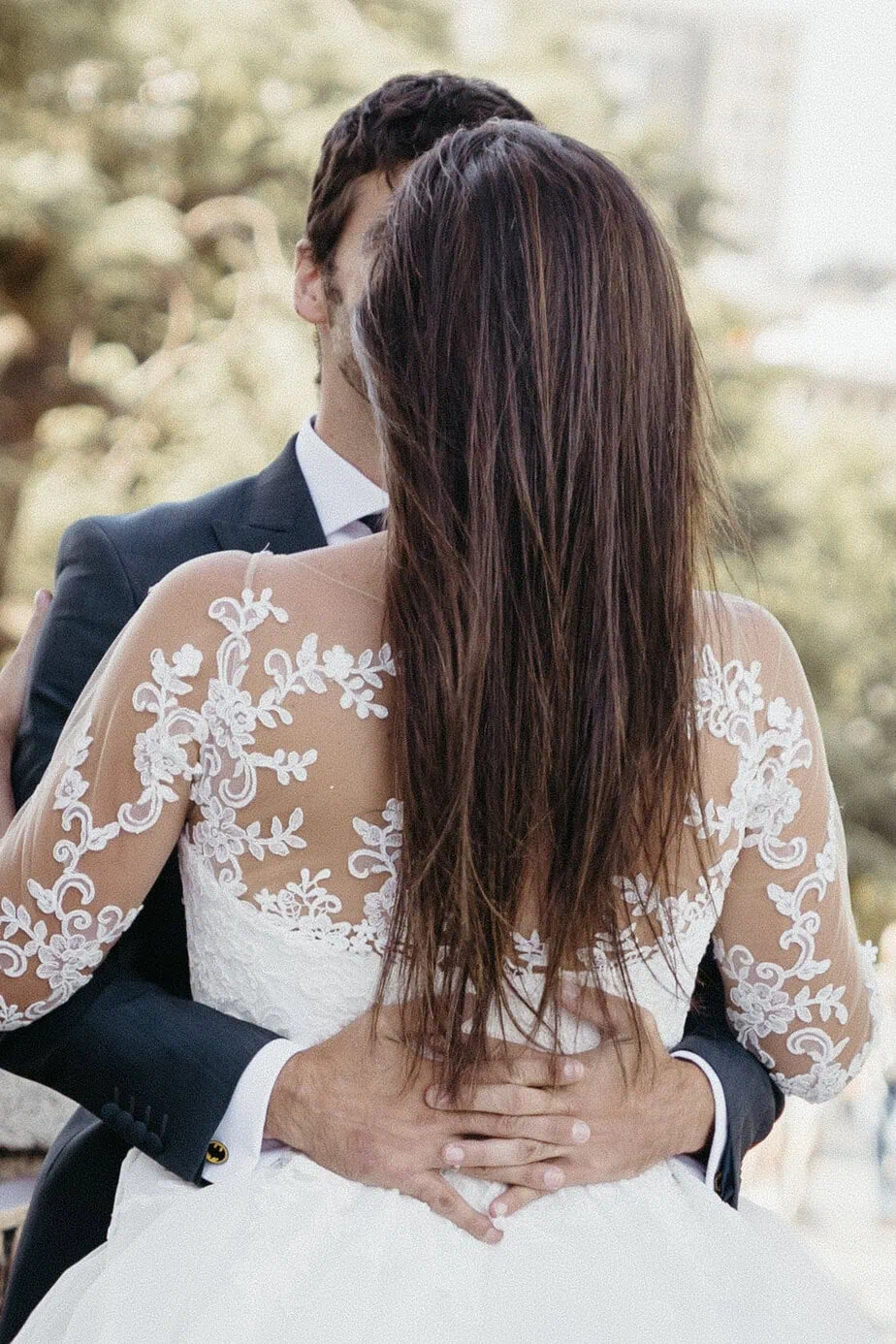 Una pareja se abraza, ella en vestido de encaje blanco y él en traje oscuro, vistos desde atrás.