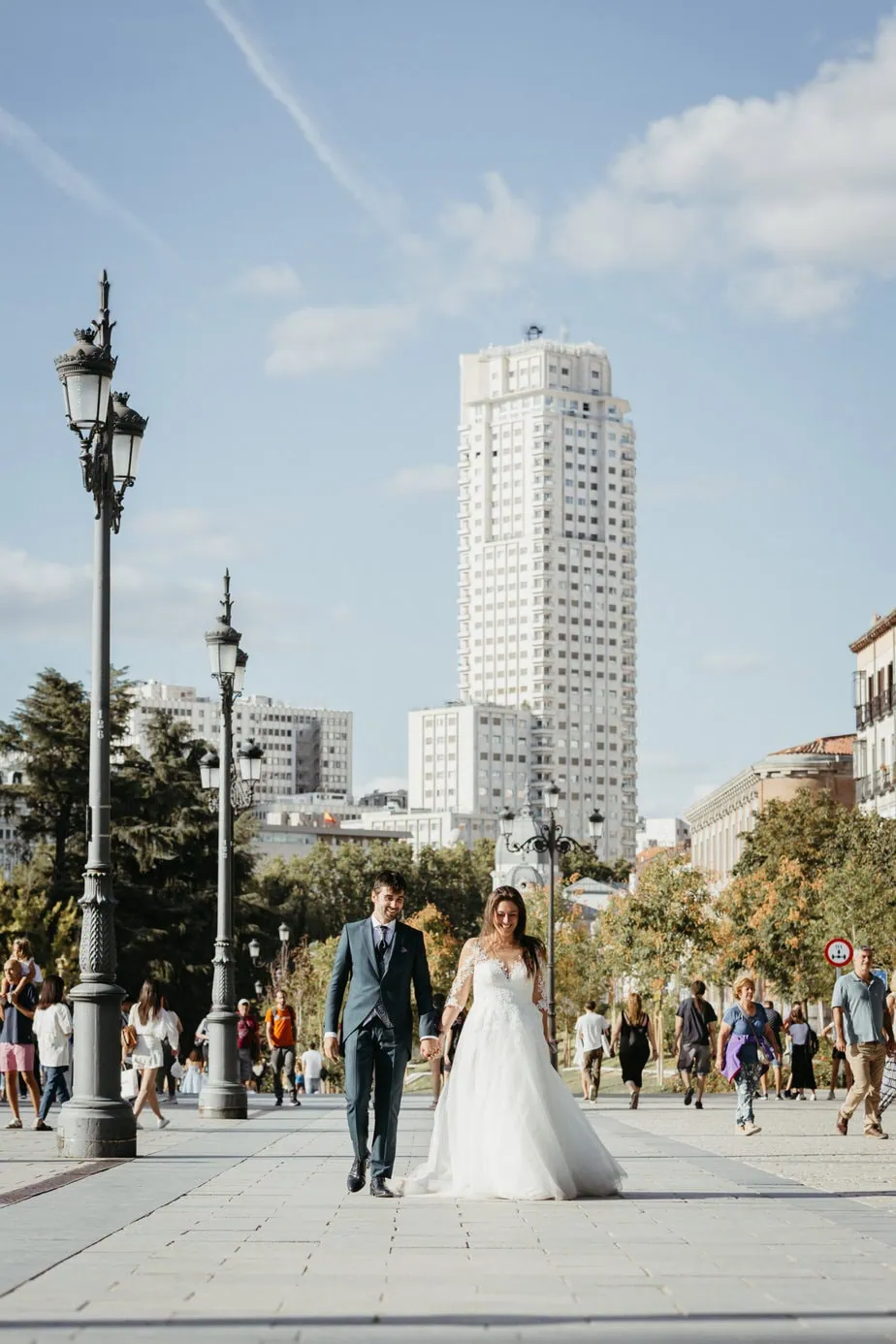 Pareja en traje y vestido de boda caminando en una avenida concurrida con un edificio al fondo.