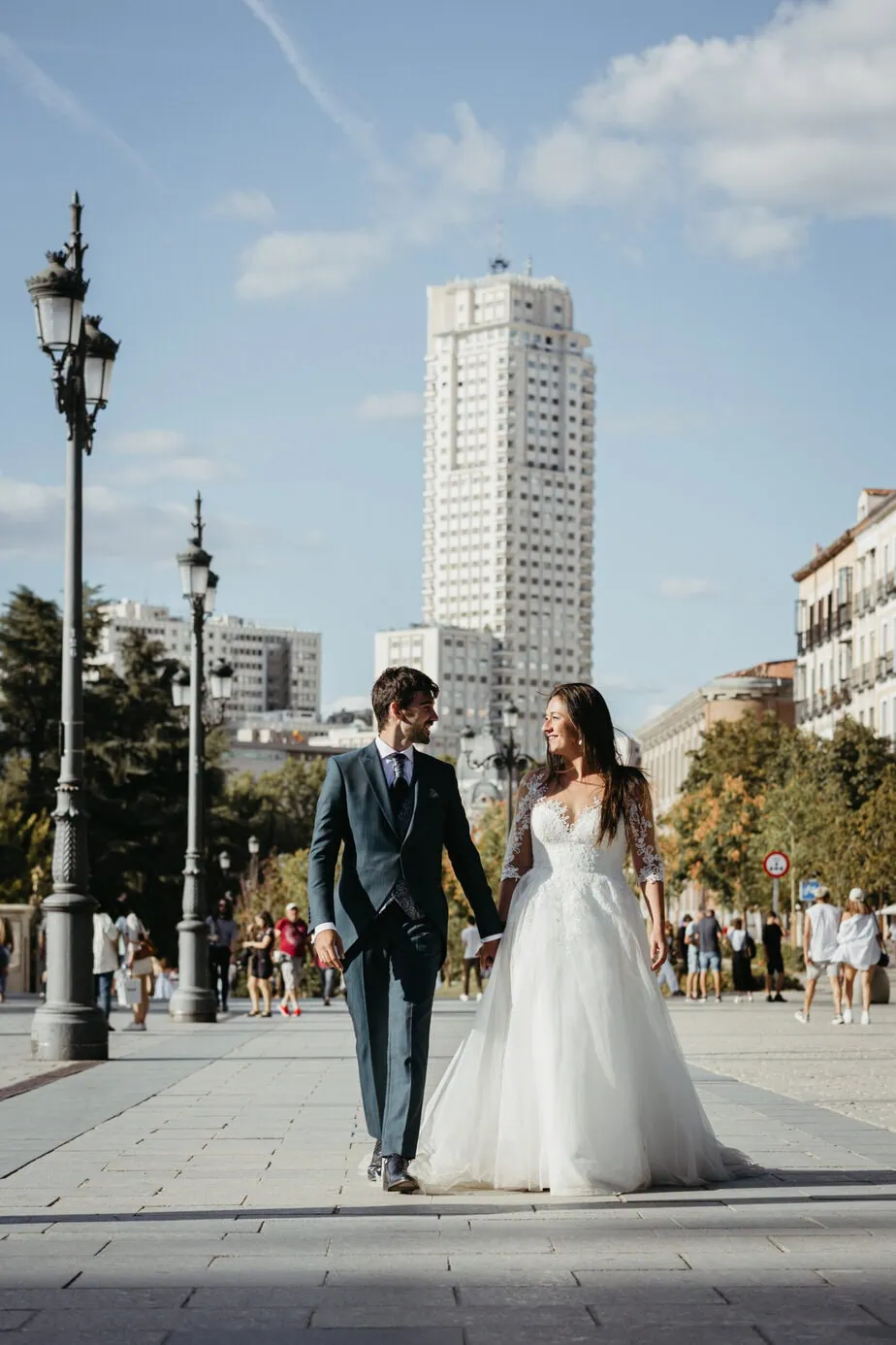 Pareja de novios caminando en una calle urbana con edificios y farolas bajo un cielo despejado.