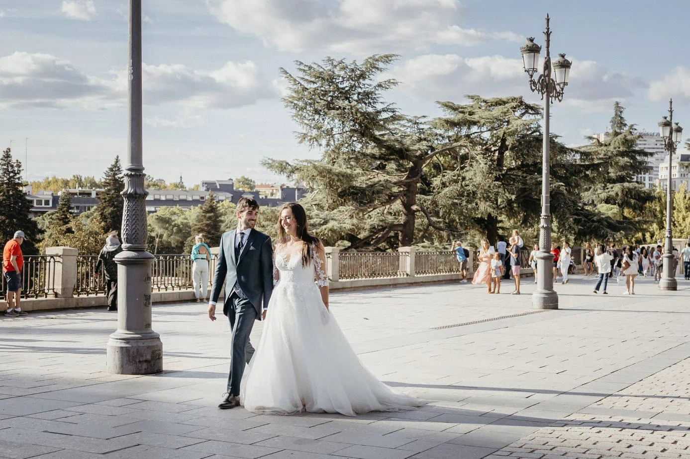 Pareja elegantemente vestida caminando en área urbana con árboles y personas paseando.