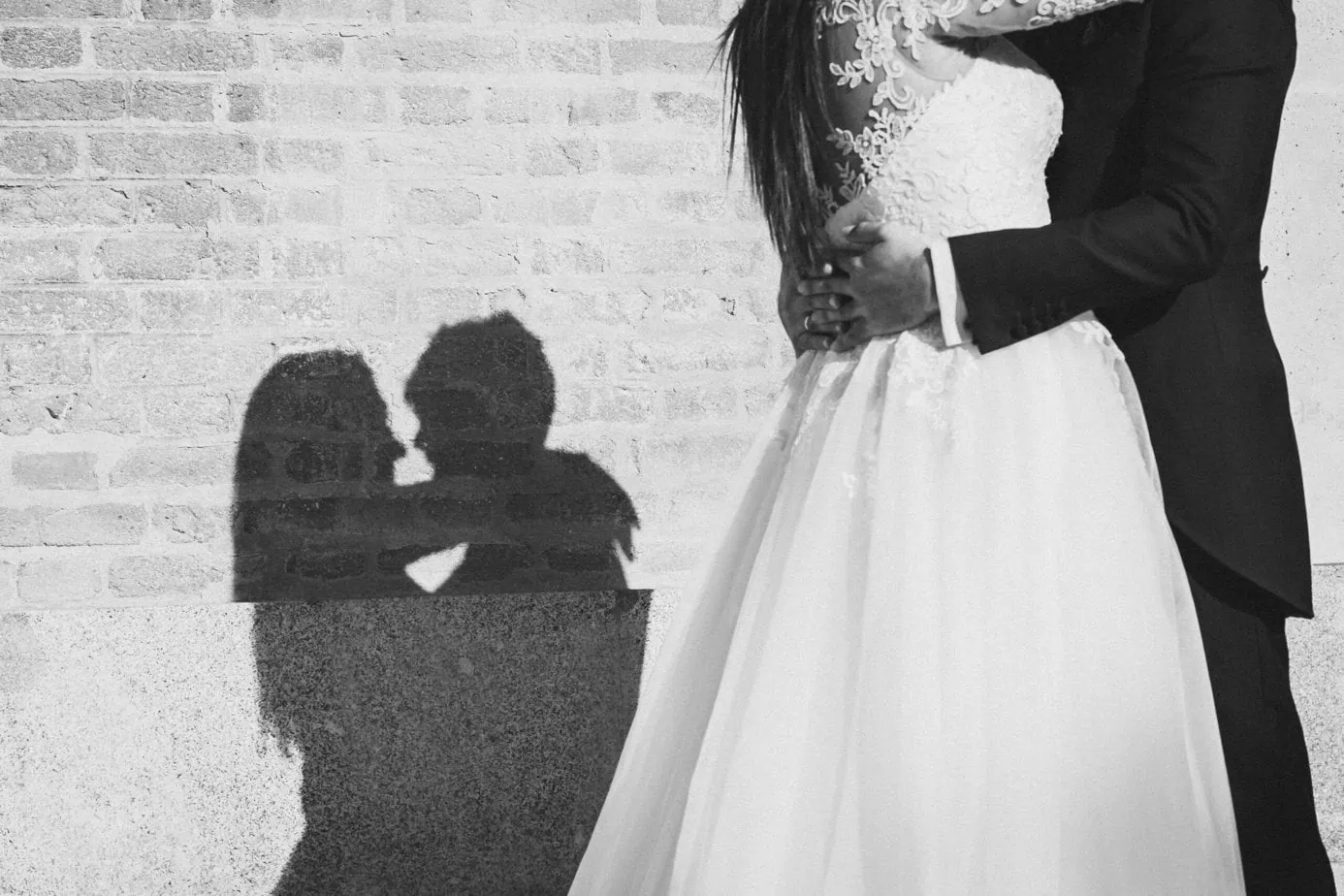 Pareja en traje de boda abrazada junto a muro, sombra proyecta un beso.