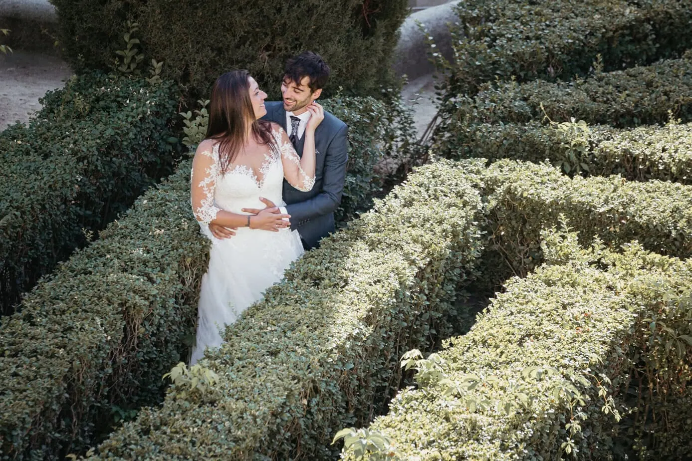 Pareja en un jardín laberinto, ella en vestido blanco y él en traje oscuro mirándose con afecto.