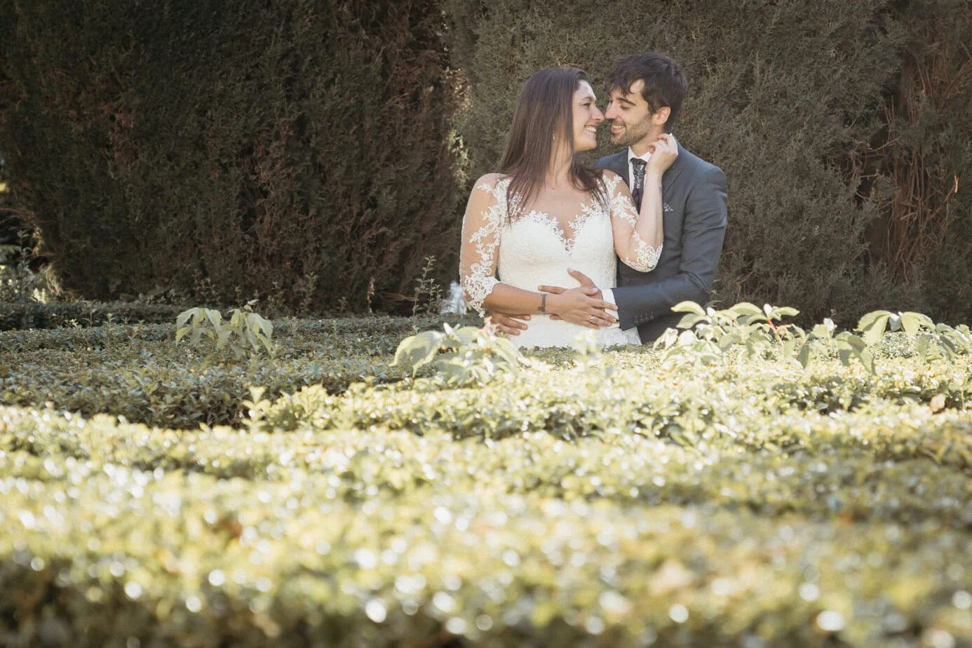 Pareja formalmente vestida abrazándose y sonriendo en un jardín con vegetación y setos altos.