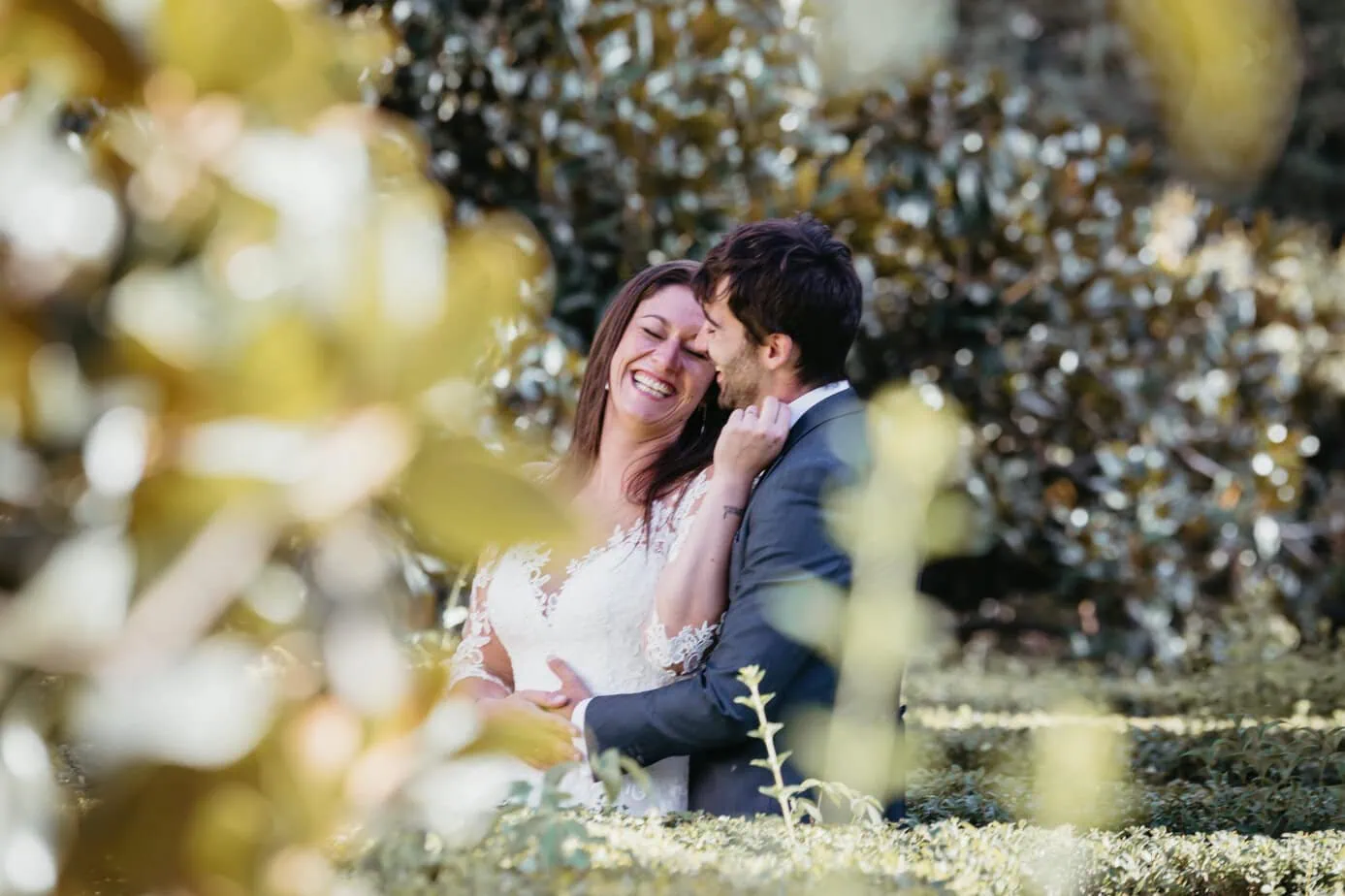 Pareja vestida de boda riendo al aire libre, rodeada de vegetación.