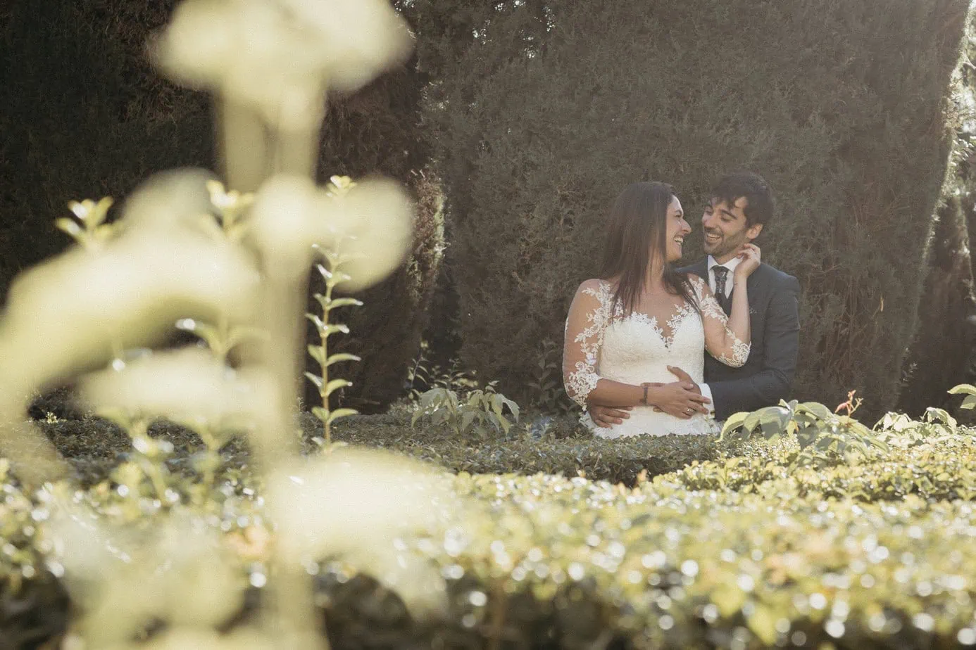 Pareja vestida de boda en jardín verde, rodeada de vegetación, se miran sonriendo.